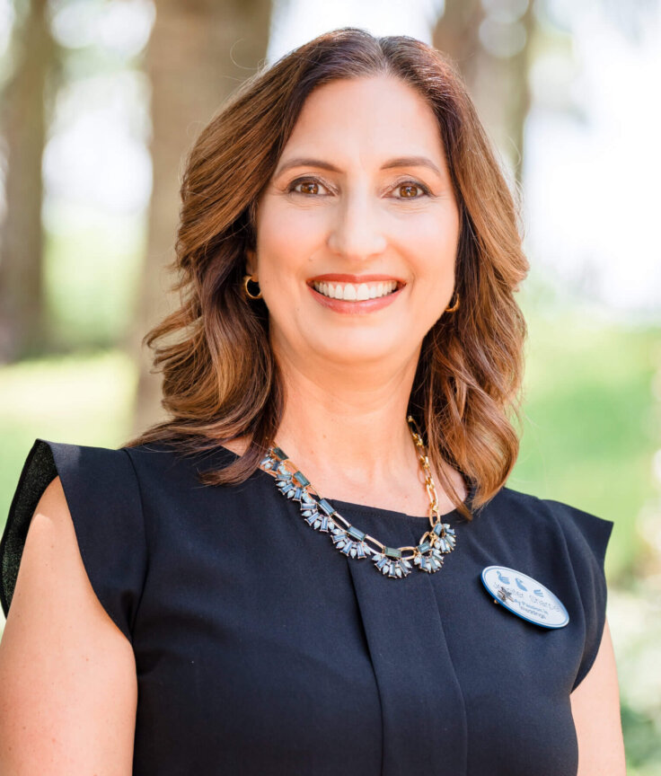 A woman with shoulder-length brown hair, wearing a black dress, a floral necklace, and a name badge, smiles outdoors with trees and greenery blurred in the background.