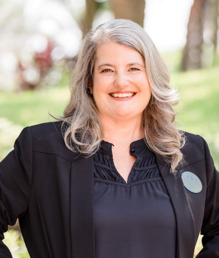A smiling woman with long gray hair wearing a black blazer and blouse stands outdoors in a sunny, green setting.