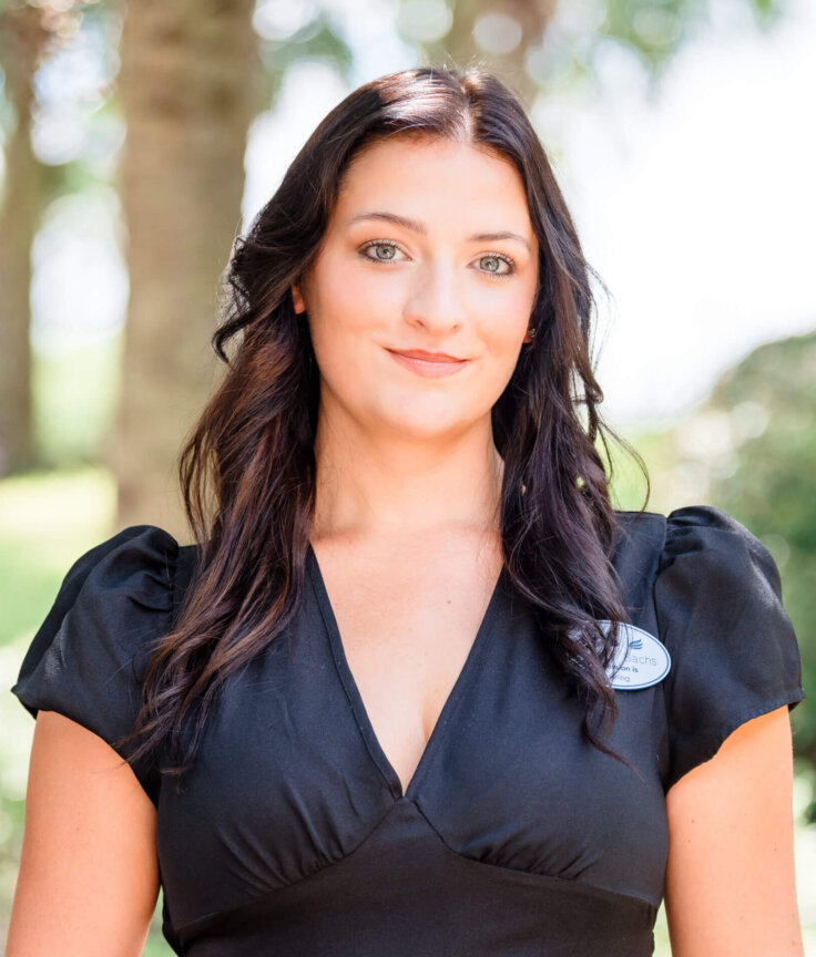 A woman with long, dark hair wearing a black V-neck dress and a name badge stands outdoors, smiling softly, with trees and greenery blurred in the background.