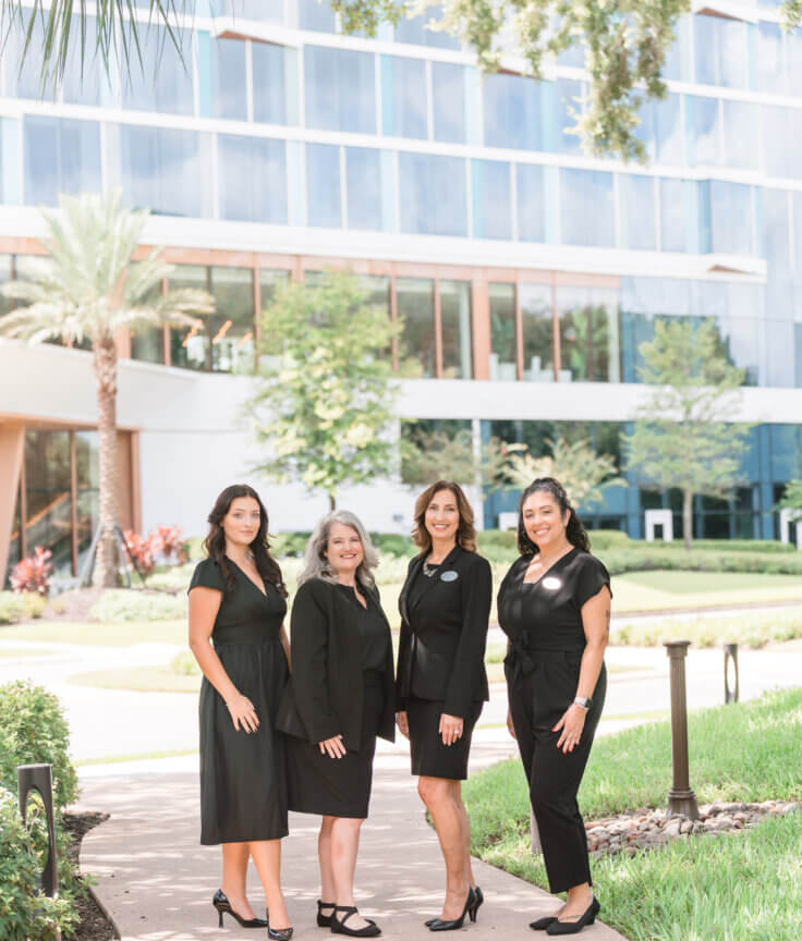 Four women in black business attire stand on a curved sidewalk outside a modern glass building, surrounded by green grass, trees, and landscaping on a sunny day.