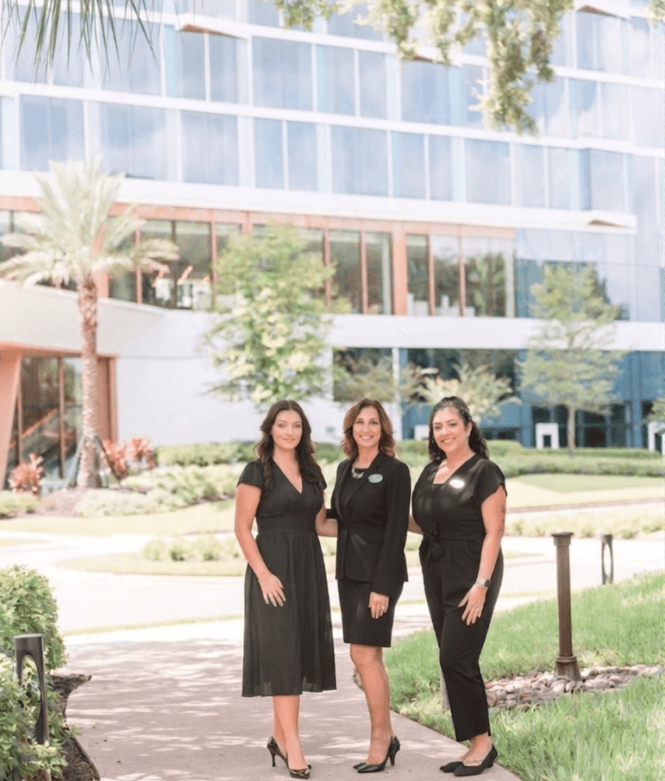 Three women wearing black business attire stand together on a curved sidewalk in front of a modern glass building, surrounded by greenery and palm trees on a sunny day.