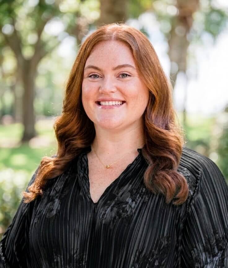 Maddie Lunn - with long red hair wearing a black blouse smiles outdoors, with sunlight and trees blurred in the background.
