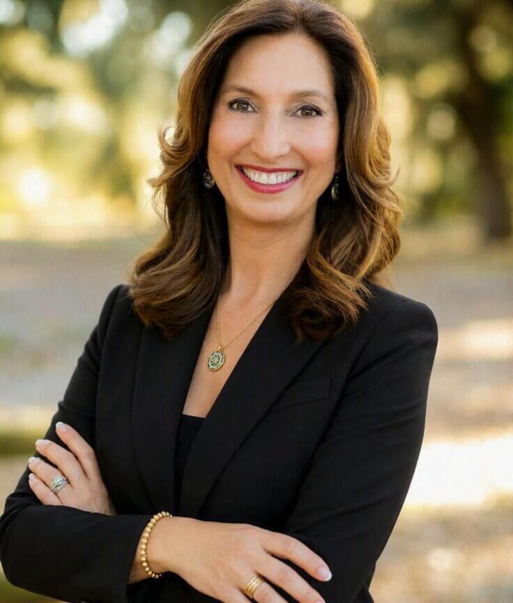 A woman with long brown hair in a black blazer smiles with arms crossed outdoors, in front of sunlit trees.